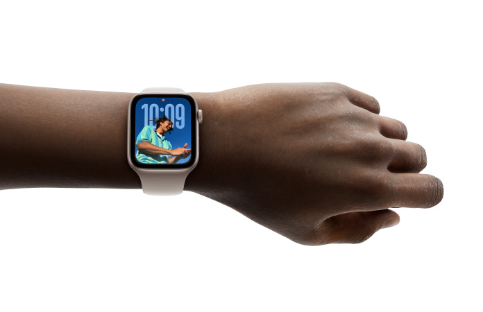 Primer plano de la muñeca de una persona con un Apple Watch en color plata y correa de goma, cuya pantalla muestra la hora en el nuevo diseño con Liquid Glass