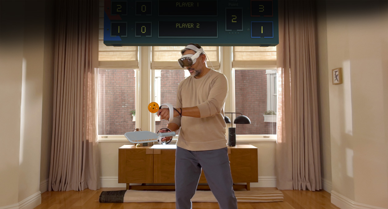 Un homme portant un Apple Vision Pro et tenant des manettes Playstation VR2 Sense Sony joue à Pickle Pro, un tableau d’affichage spatial au-dessus de la tête